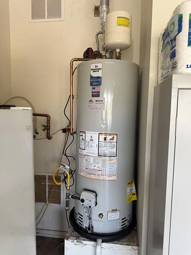 A Bradford White gas water heater on a raised stand with an expansion tank above it, copper supply lines and a Capstone service sticker on the front