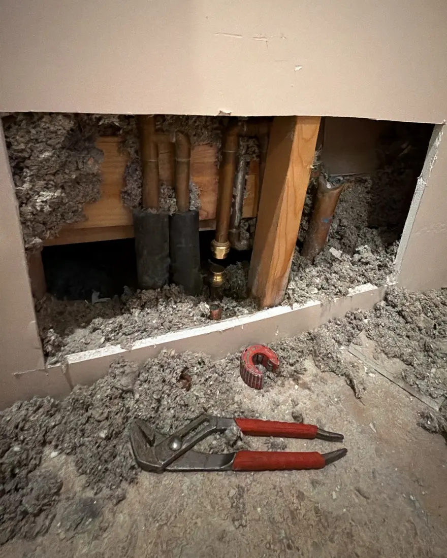 Copper pipes with a new brass fitting inside an opened wall during a pipe repair, with tools staged below