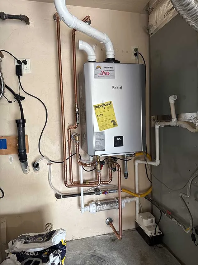 Rinnai tankless water heater with a Capstone Plumbing sticker, mounted on a utility wall with white PVC vent pipes above and copper water lines, a yellow gas line and white condensate piping below