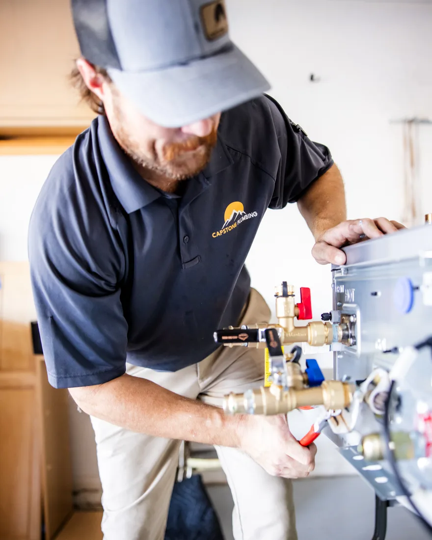 A Capstone plumber in a branded polo fitting the manifold and service valves on a wall-mounted tankless water heater