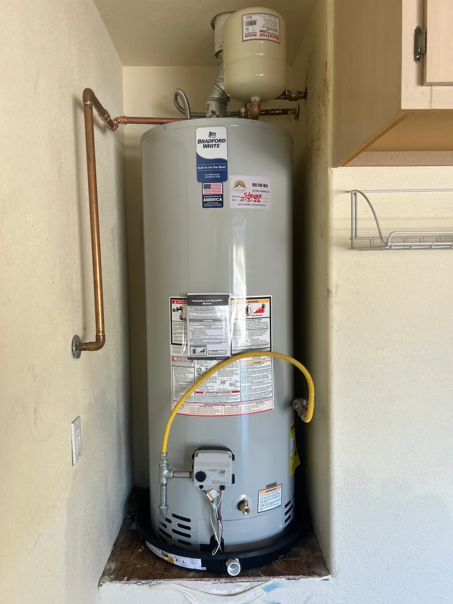 A newly installed Bradford White gas water heater with expansion tank above and a dated Capstone service tag on the front