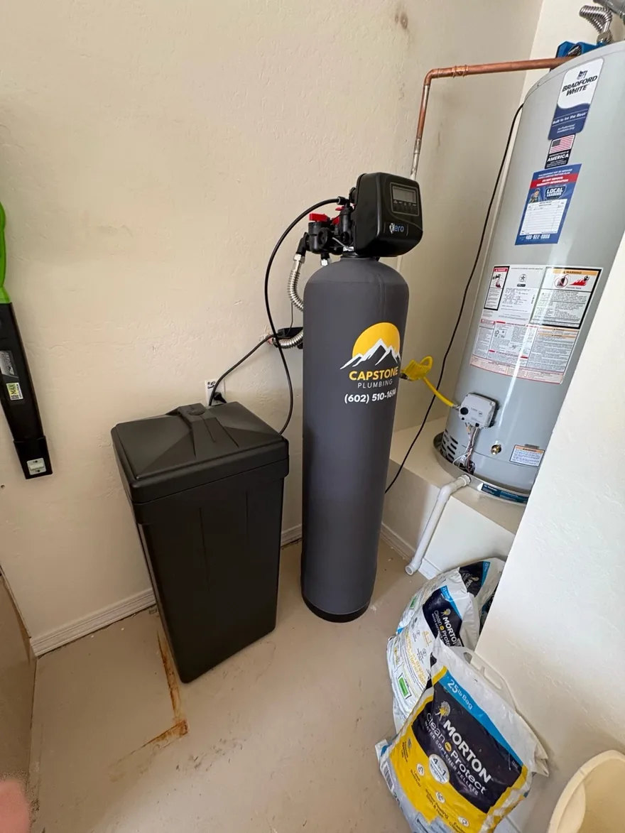 Water softener in a gray Capstone Plumbing insulating jacket beside its brine tank and a Bradford White water heater in a garage