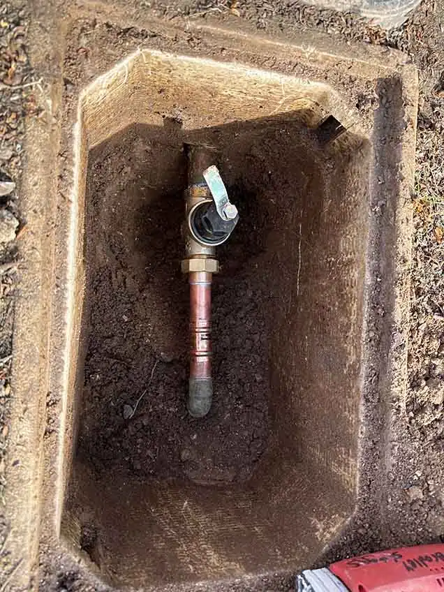 A new copper shutoff valve with a lever handle on a copper riser, exposed inside a dug-out in-ground valve box