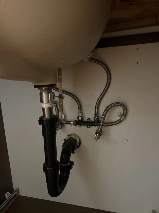 The underside of a sink showing braided stainless supply lines, a shutoff valve and a black P-trap, with the cabinet dry